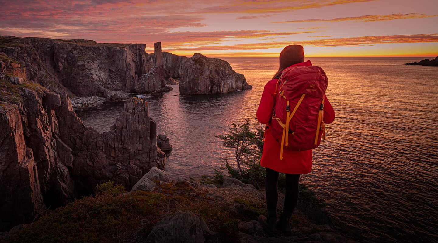 Person wearing a red jacket and large backpack stands on a rocky cliff, overlooking the ocean and dramatic coastline at sunset.