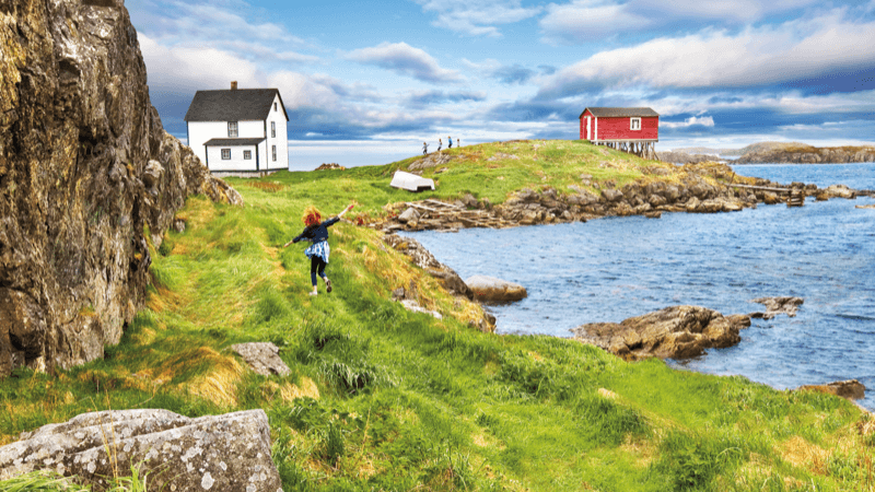 A young girl is playfully running across the green landscape of Change Islands. She is heading towards a white house and red fishing shed.