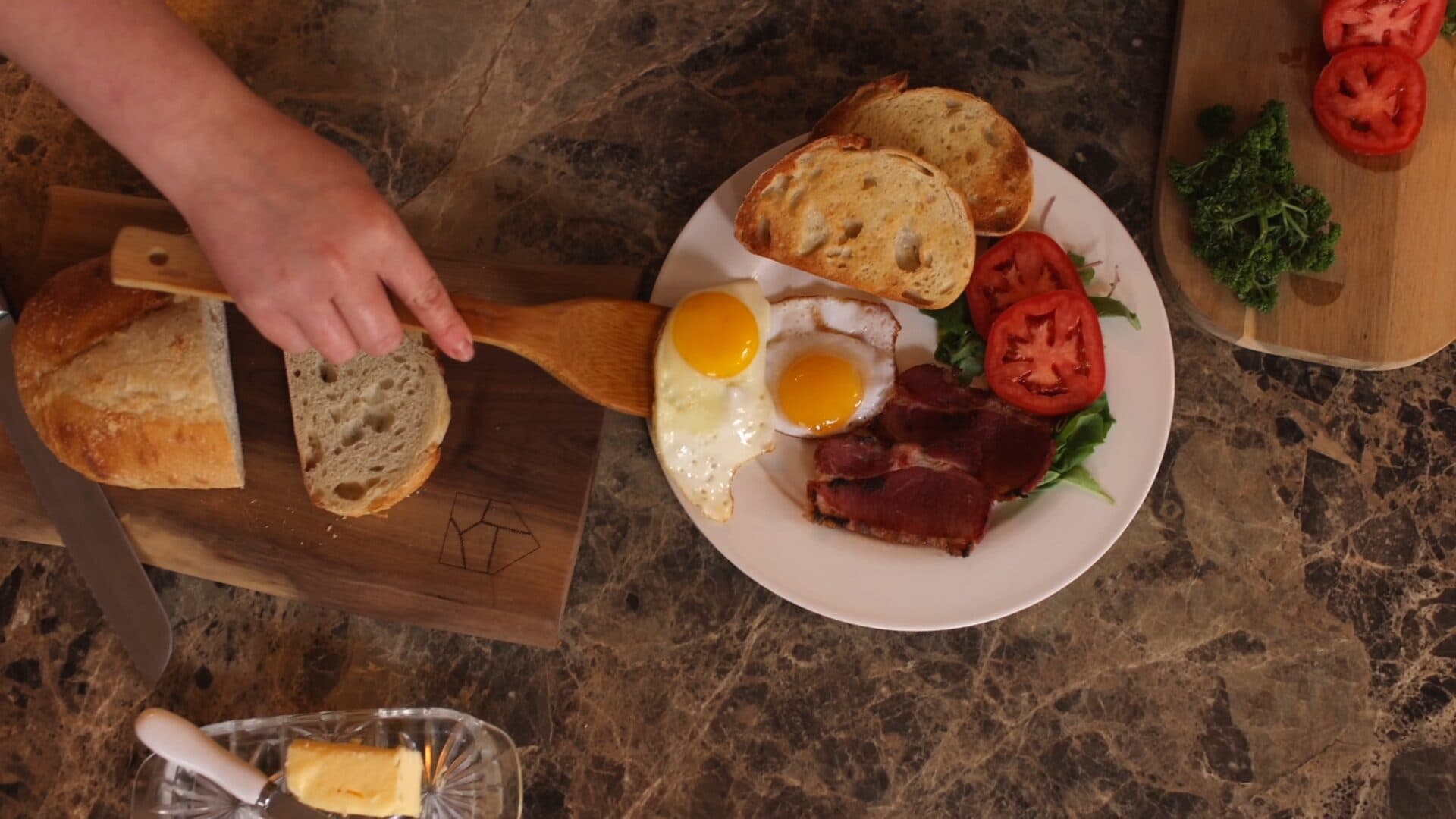 A person places slices of bread on a cutting board next to a plate with two sunny-side-up eggs, bacon, toast, tomato slices, and greens on a marble surface. Butter and more tomatoes are also visible.