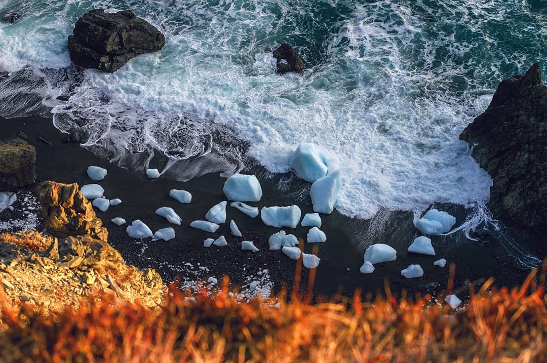 Aerial view of a rocky coastline with waves crashing onto the shore. Large white ice pieces are scattered along the dark sandy beach. The foreground shows vibrant reddish-brown foliage, contrasting with the dark rocks and blue-green ocean.