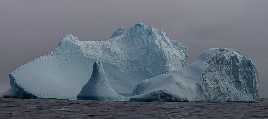 A large iceberg with rugged, jagged peaks floats in dark, calm waters under an overcast sky. The ice displays various shades of blue and white, with smooth and craggy textures. The scene conveys a sense of cold and isolation.