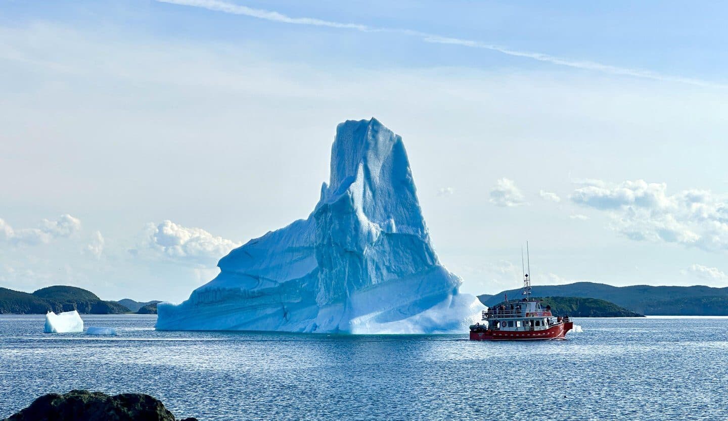 A large iceberg floats in the ocean with a small red and white boat nearby. Rocky shores and distant islands are visible under a blue sky with scattered clouds.