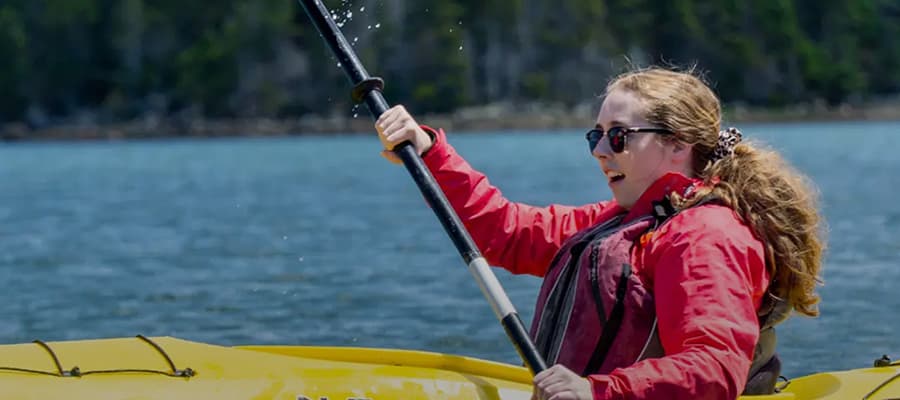 A person wearing a red jacket and sunglasses is kayaking on a body of water. They hold a paddle and appear to be enjoying the activity. The background features a wooded shoreline.