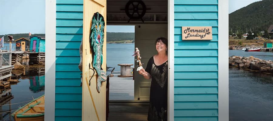 A scenic coastal village is bathed in warm sunset light. Colorful houses are nestled among lush green hills, with vibrant purple flowers in the foreground. The calm sea and misty islands are visible in the background.