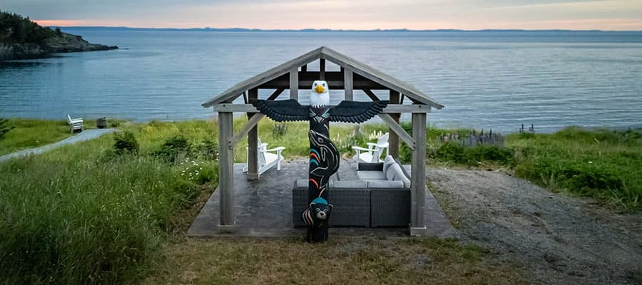 A person wearing a red jacket and sunglasses is kayaking on a body of water. They hold a paddle and appear to be enjoying the activity. The background features a wooded shoreline.