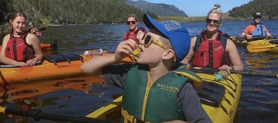 A group of people kayaking on a lake, surrounded by lush green forests and mountains. A boy in the foreground is wearing a life jacket and sunglasses, blowing a whistle while seated in a yellow kayak. Others smile in the background.