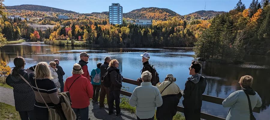 A group of people kayaking on a lake, surrounded by lush green forests and mountains. A boy in the foreground is wearing a life jacket and sunglasses, blowing a whistle while seated in a yellow kayak. Others smile in the background.