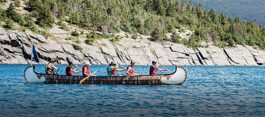 A red bicycle is parked on a paved path beside a body of water. The bike has wide tires, a rear rack, and a curvy frame. The surrounding area has gravel and sparse grass near the water’s edge.