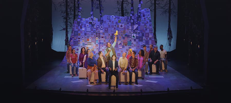 A colorful stage set shows eleven people seated in a row, surrounded by a backdrop filled with numerous square screens displaying various icons. The background is designed to resemble a forest with tree silhouettes.