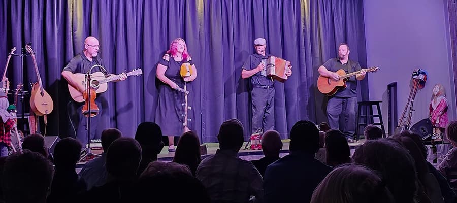 Four musicians perform on stage against a dark backdrop. The group consists of two guitarists, a bagpiper, and a person playing an accordion. The audience is seated in front, watching the performance. Musical instruments are visible to the side.