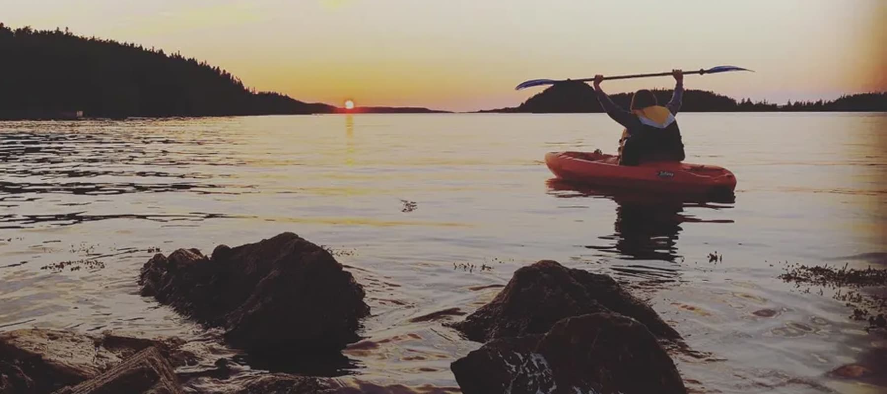 A person in a kayak raises a paddle triumphantly while sitting on calm water. The sun sets on the horizon, casting an orange glow over the scene. Rocky shores and silhouetted trees frame the serene landscape.