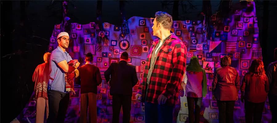 Four musicians perform on stage against a dark backdrop. The group consists of two guitarists, a bagpiper, and a person playing an accordion. The audience is seated in front, watching the performance. Musical instruments are visible to the side.