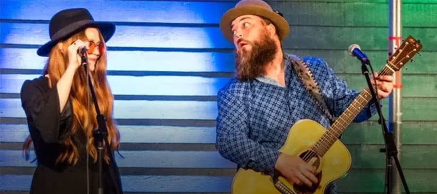 A person in a kayak raises a paddle triumphantly while sitting on calm water. The sun sets on the horizon, casting an orange glow over the scene. Rocky shores and silhouetted trees frame the serene landscape.