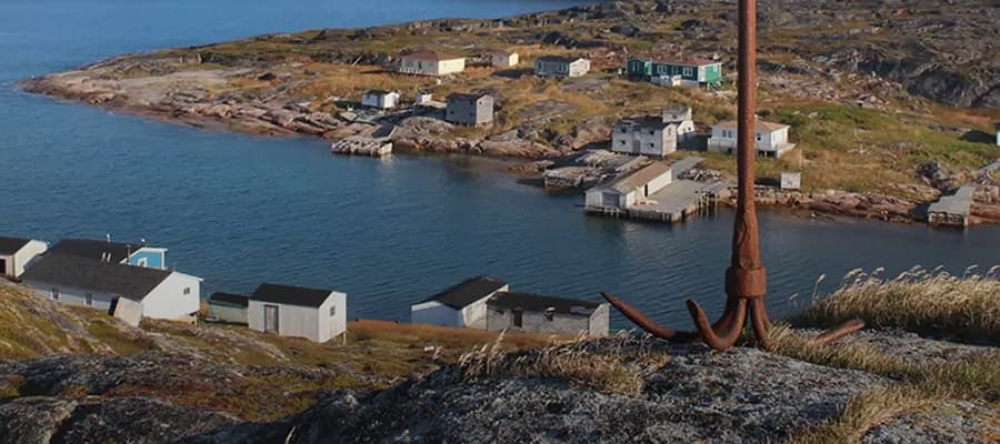 Small coastal village with several houses scattered along rocky terrain by the sea. An old rusted anchor is prominent in the foreground. The landscape features rugged rocks and sparse vegetation under a clear blue sky.