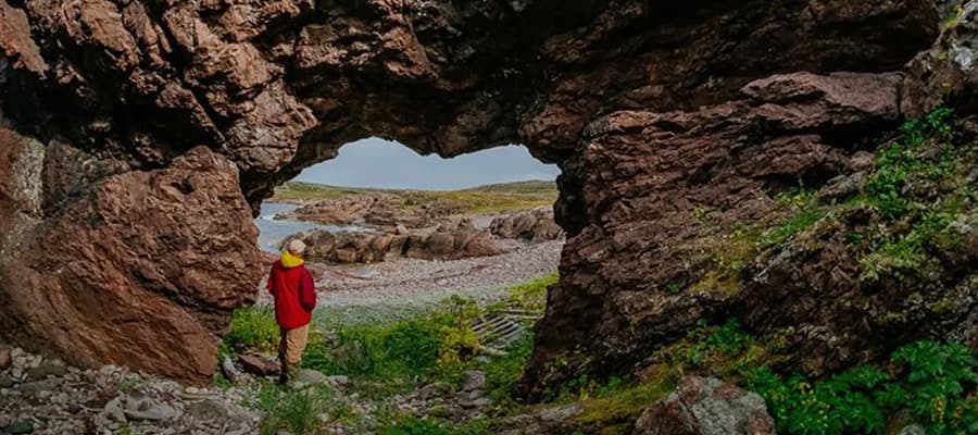 A person in a red jacket and yellow hat stands facing a rocky arch with greenery. They overlook a rugged landscape with distant water and rocks under a cloudy sky.