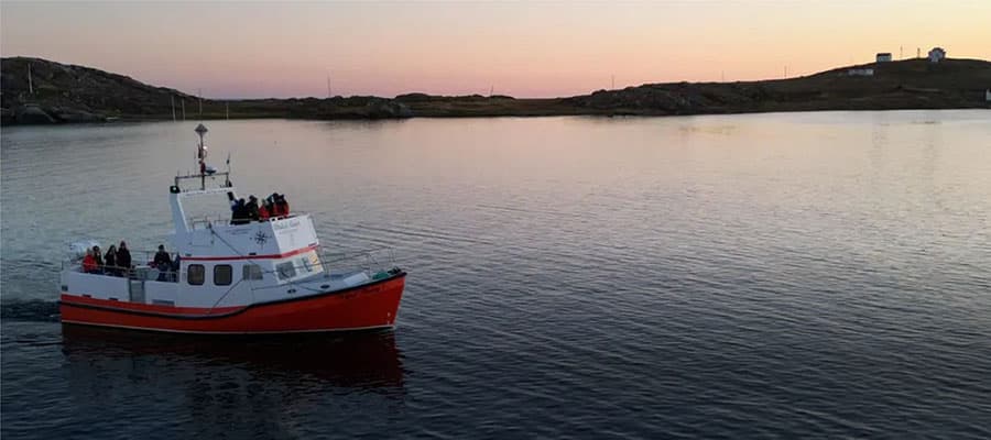 A person in a red jacket and yellow hat stands facing a rocky arch with greenery. They overlook a rugged landscape with distant water and rocks under a cloudy sky.