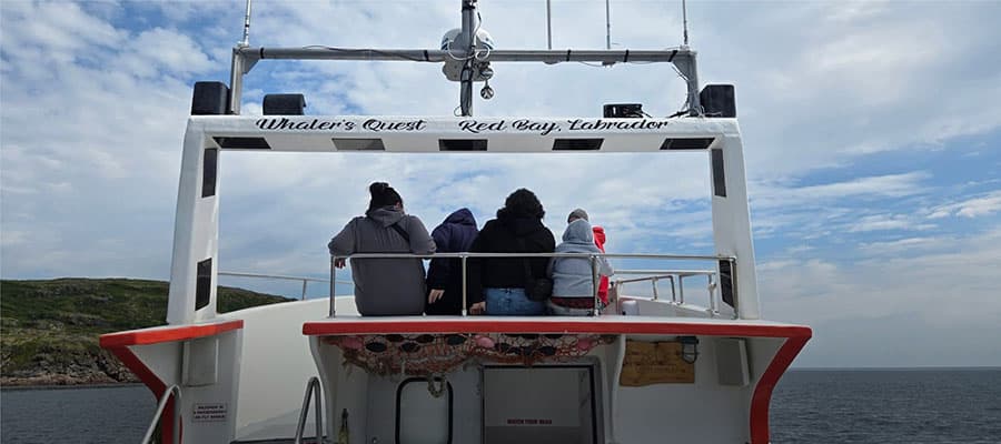 Small coastal village with several houses scattered along rocky terrain by the sea. An old rusted anchor is prominent in the foreground. The landscape features rugged rocks and sparse vegetation under a clear blue sky.