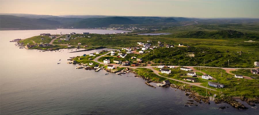 Two people are walking across a grassy landscape near the rocky coast of a large body of water. A hilly terrain with patches of vegetation surrounds them under a partly cloudy sky.