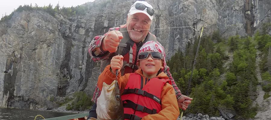 A smiling elderly man and a child, both wearing life vests, proudly holding a fish on a boat. Behind them, there's a scenic view of rocky cliffs and green trees. The child is wearing a bandana and sunglasses.