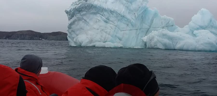 People in red jackets in a small boat observe a large, jagged iceberg floating on calm, dark waters. In the distance, rocky coastline is visible under an overcast sky.