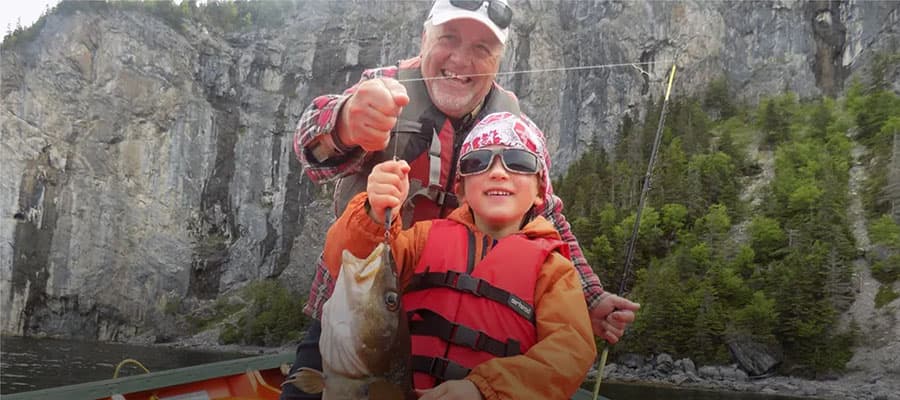A smiling elderly man and a child, both wearing life vests, proudly holding a fish on a boat. Behind them, there's a scenic view of rocky cliffs and green trees. The child is wearing a bandana and sunglasses.