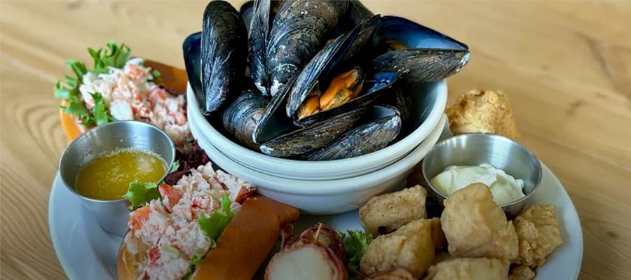 People in red jackets in a small boat observe a large, jagged iceberg floating on calm, dark waters. In the distance, rocky coastline is visible under an overcast sky.