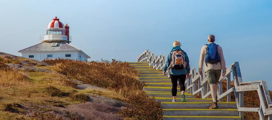 Two people wearing backpacks walk up wooden stairs towards a lighthouse with a red dome. The sky is clear, and the surrounding landscape is grassy with shrubs.