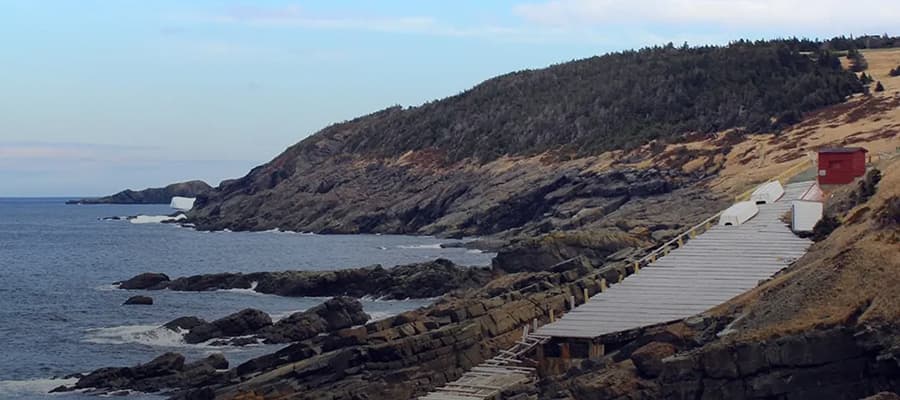 A rocky coastal landscape with a narrow wooden pathway leading along the shore. Cliffs covered with sparse vegetation rise to the right. The ocean is calm, with a few small icebergs visible in the water. A red building stands near the path.