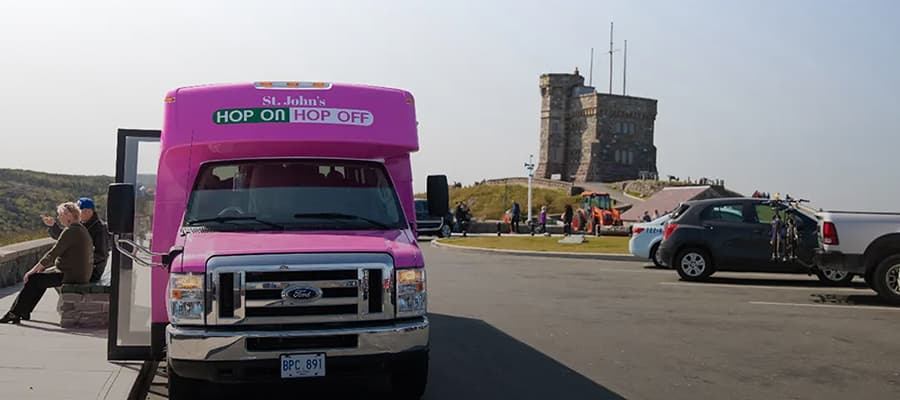 Pink tour bus parked at a scenic spot with "St. John's Hop On Hop Off" written on it. A few people sit on a bench nearby, and a historic stone building stands in the background under a clear sky.