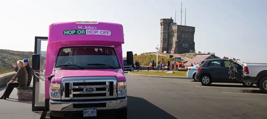 A rocky coastal landscape with a narrow wooden pathway leading along the shore. Cliffs covered with sparse vegetation rise to the right. The ocean is calm, with a few small icebergs visible in the water. A red building stands near the path.