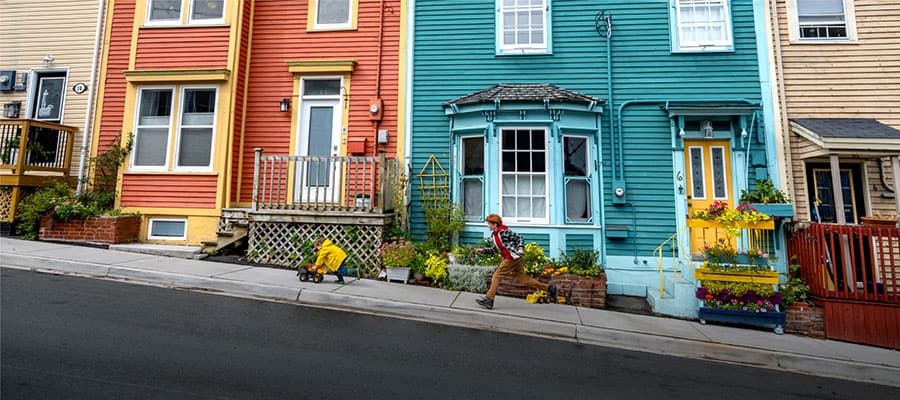 Pink tour bus parked at a scenic spot with "St. John's Hop On Hop Off" written on it. A few people sit on a bench nearby, and a historic stone building stands in the background under a clear sky.