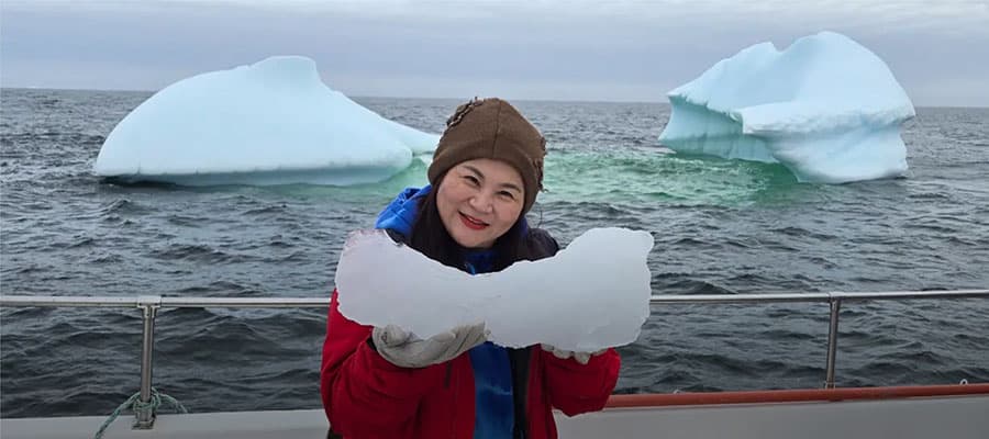 A person in a red jacket and yellow hat stands under a large, rocky arch looking out at a scenic landscape. The view includes grassy terrain, rocky ground, and a distant body of water under an overcast sky.