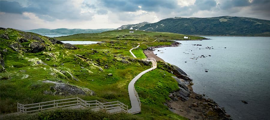Two people walk along a grassy, rocky coastline with a clear blue sky overhead. The ocean is to their right, with waves lapping against the shore. The terrain is rugged, with patches of shrubs and hills in the background.