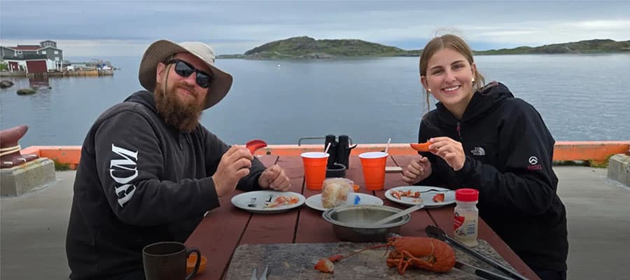 A small red and white boat with passengers travels across calm water at sunset. The sky is a gradient of warm colors, and the silhouette of a hilly shoreline is visible in the background.