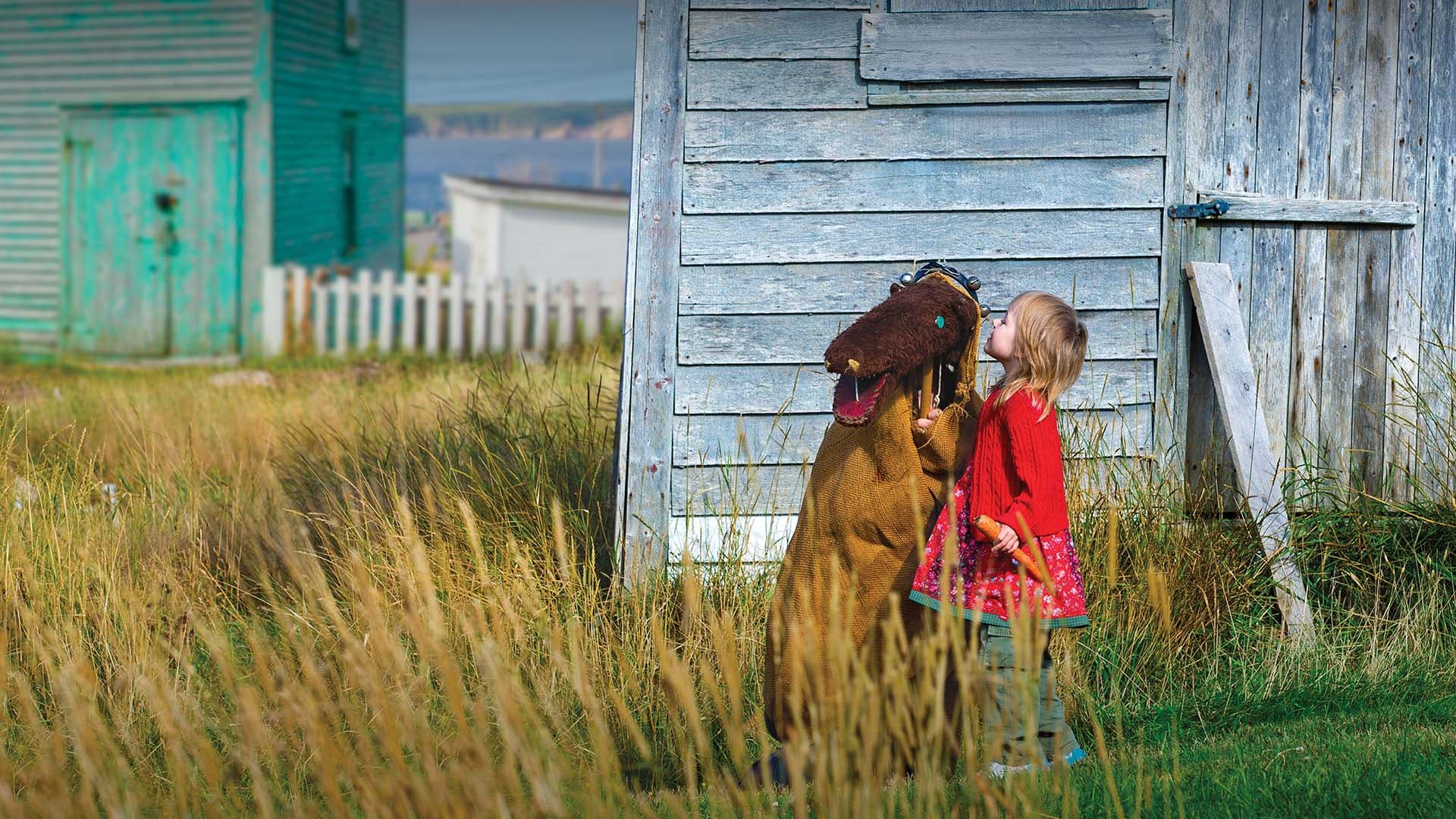 A young girl in a red dress interacts with a hobby horse beside a weathered wooden shed.