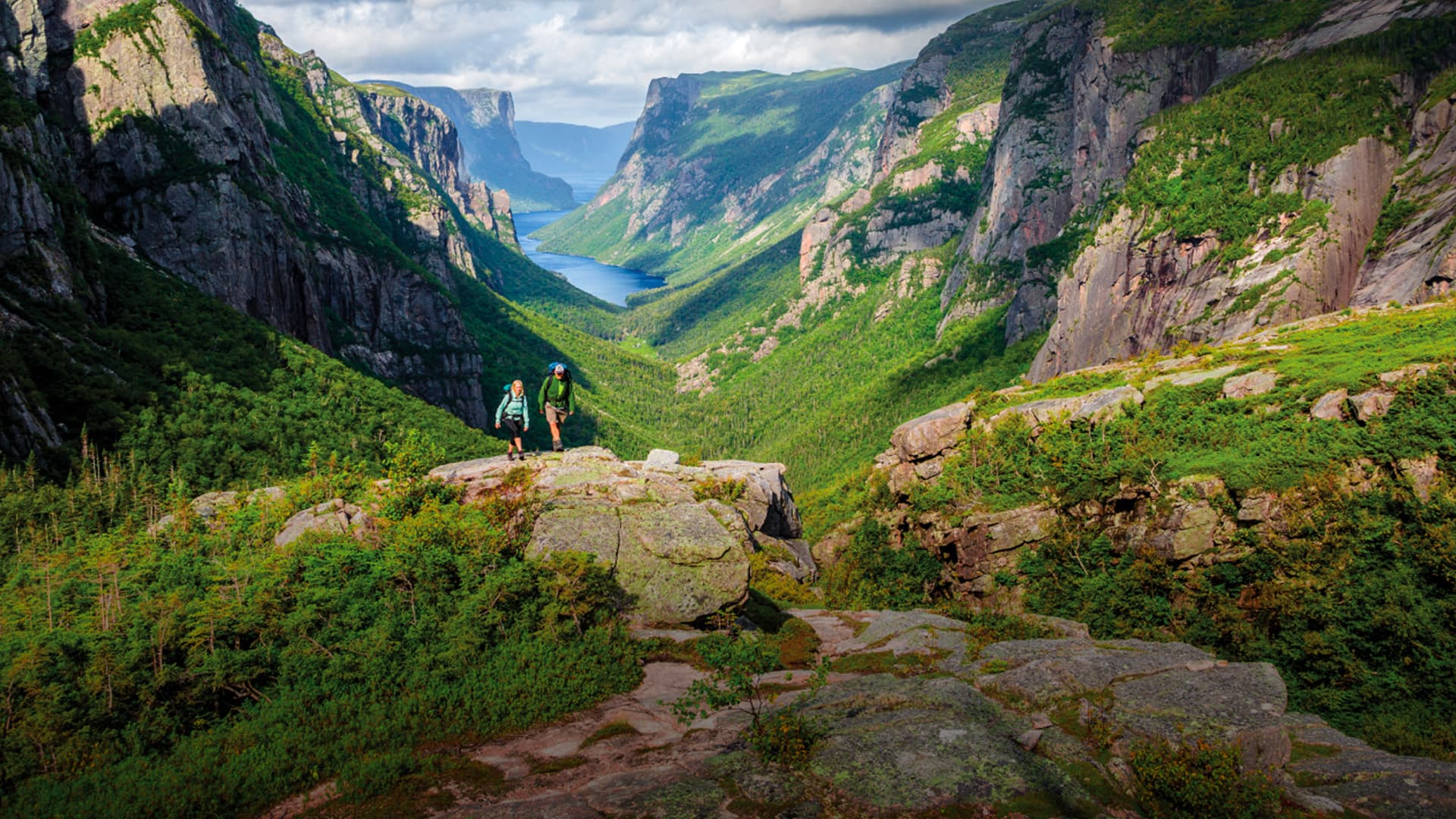 Two hikers stand on a rocky outcrop overlooking a lush, green valley with steep cliffs.