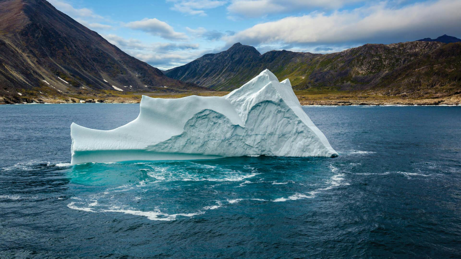 A massive iceberg is floating by an ancient mountain range.