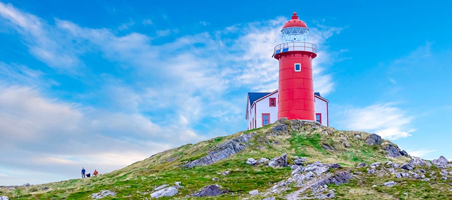 Hikers are walking up the rugged hills toward a red lighthouse in Ferryland.