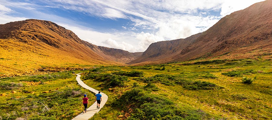 Two travellers are hiking on a path toward the red landscape of the Tablelands in Gros Morne National Park.