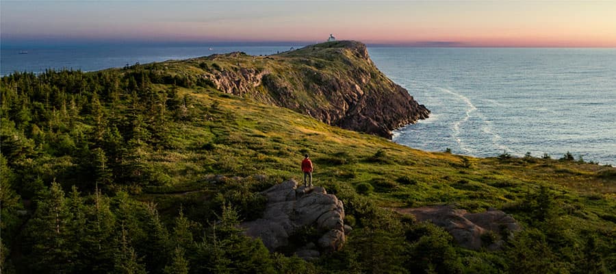 Three people on a boat, two men and a woman, are fishing. One man, wearing a red jacket and sunglasses, is holding a fishing rod with a fish on the line, smiling. The others are standing and sitting, also smiling. The sky is cloudy.