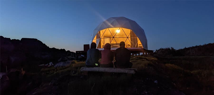 Three people walking along a grassy path beside a small coastal village with houses scattered by the water. The sky is clear and blue.