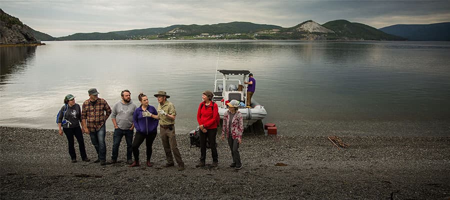 A group of people stand on a rocky shoreline near a calm body of water with hills in the background. A boat is docked behind them, and one person is seated in it. The sky is overcast, creating a muted, serene atmosphere.