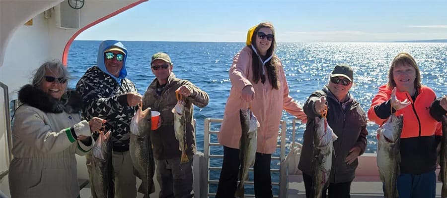 A group of people posing together outside on a wooden deck in front of a white house. They are dressed casually, and two guitars are visible at the front of the group. The sun is shining brightly.