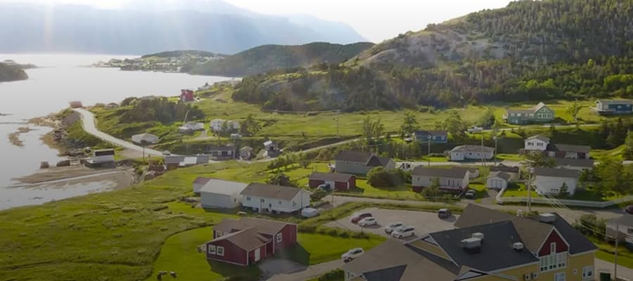 A scenic aerial view of a coastal village with scattered houses and lush greenery. A winding road follows the shoreline, while hills rise in the background. Sunlight filters through clouds, adding a warm glow to the serene landscape.