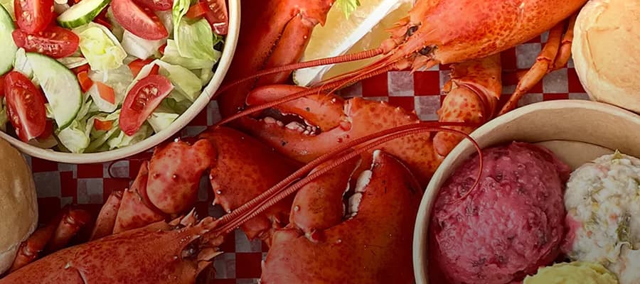 A cooked lobster with its claws spread out is alongside bowls of salad, a bread roll, coleslaw, and beet salad on a red checkered background.