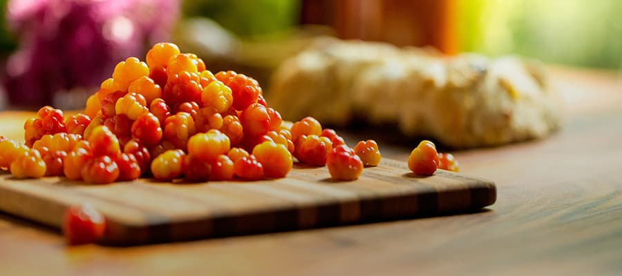 A pile of fresh cloudberries sits on a wooden cutting board. In the background, there's blurred imagery of green foliage and a wooden table. The cloudberries are vibrant orange and red, indicating ripeness.