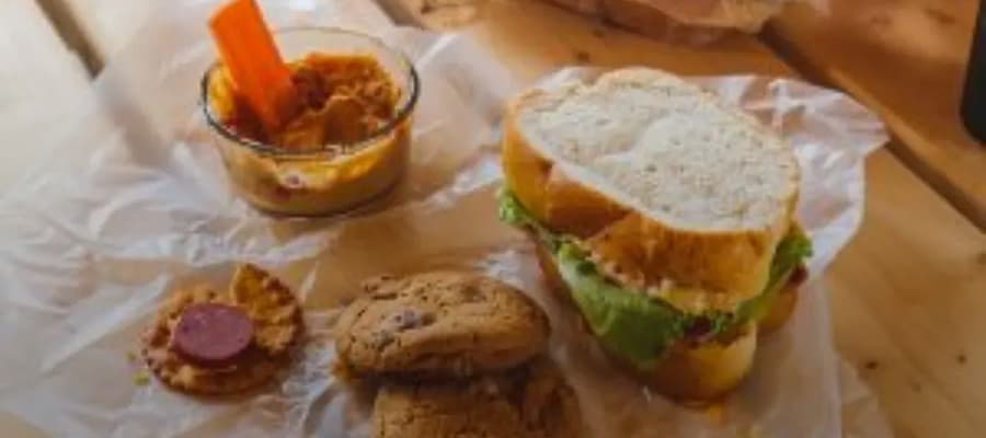 A sandwich with lettuce and filling on paper, a small bowl of hummus with a carrot stick, two cookies, and a cracker with a slice of sausage are displayed on a wooden surface.