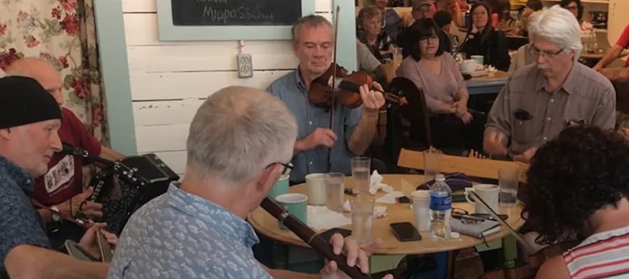 A group of five men plays traditional music with instruments including a violin, accordion, and flute at a small venue. The audience sits around enjoying the performance. The setting is cozy, with wooden decor and a chalkboard on the wall.
