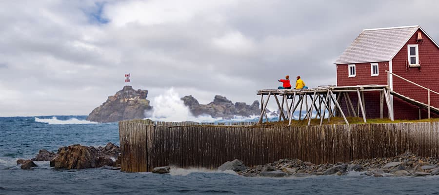 A scenic aerial view of a coastal village with scattered houses and lush greenery. A winding road follows the shoreline, while hills rise in the background. Sunlight filters through clouds, adding a warm glow to the serene landscape.