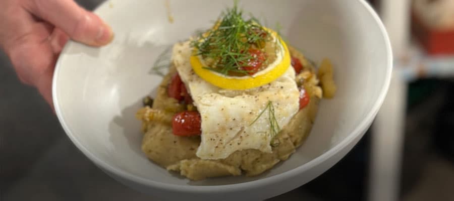 A cooked lobster with its claws spread out is alongside bowls of salad, a bread roll, coleslaw, and beet salad on a red checkered background.